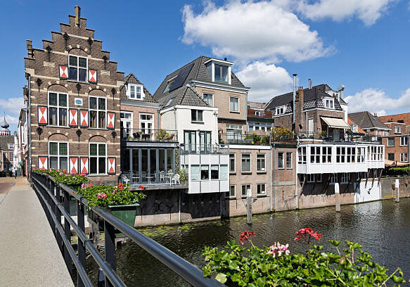 Charming traditional Dutch houses line the historic canal in Gorinchem, adorned with balconies and reflective waters, under a bright blue sky.